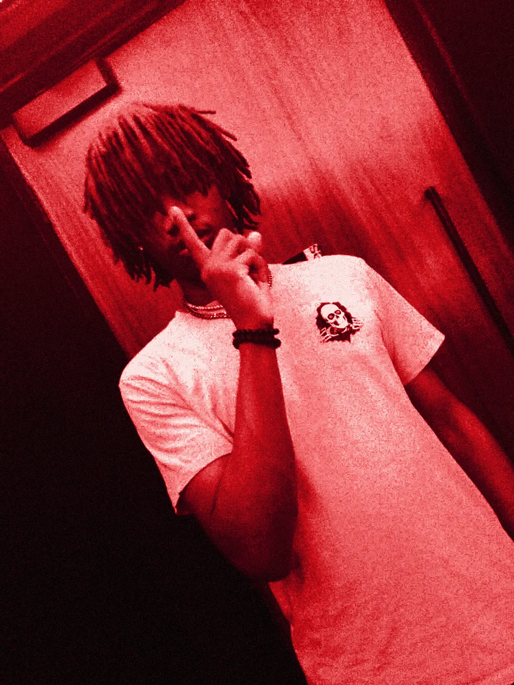 yukan in a white tee in a room flooded with red light, two fingers raised over his face.
