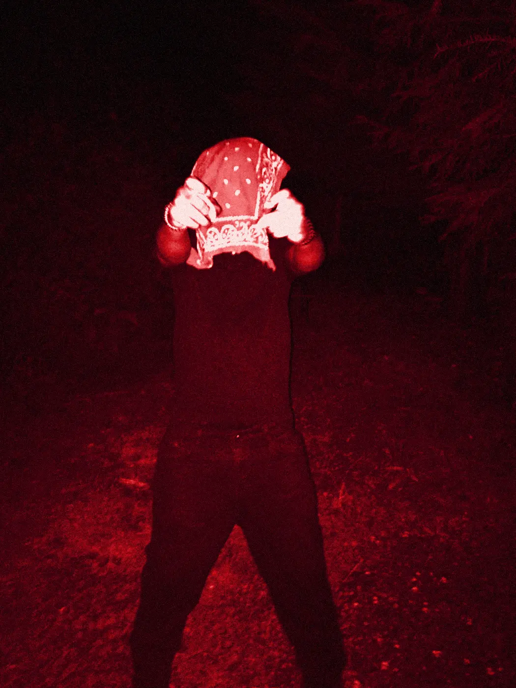 A figure holding a bandana over his face in a dark garden, blown out by the flash.
