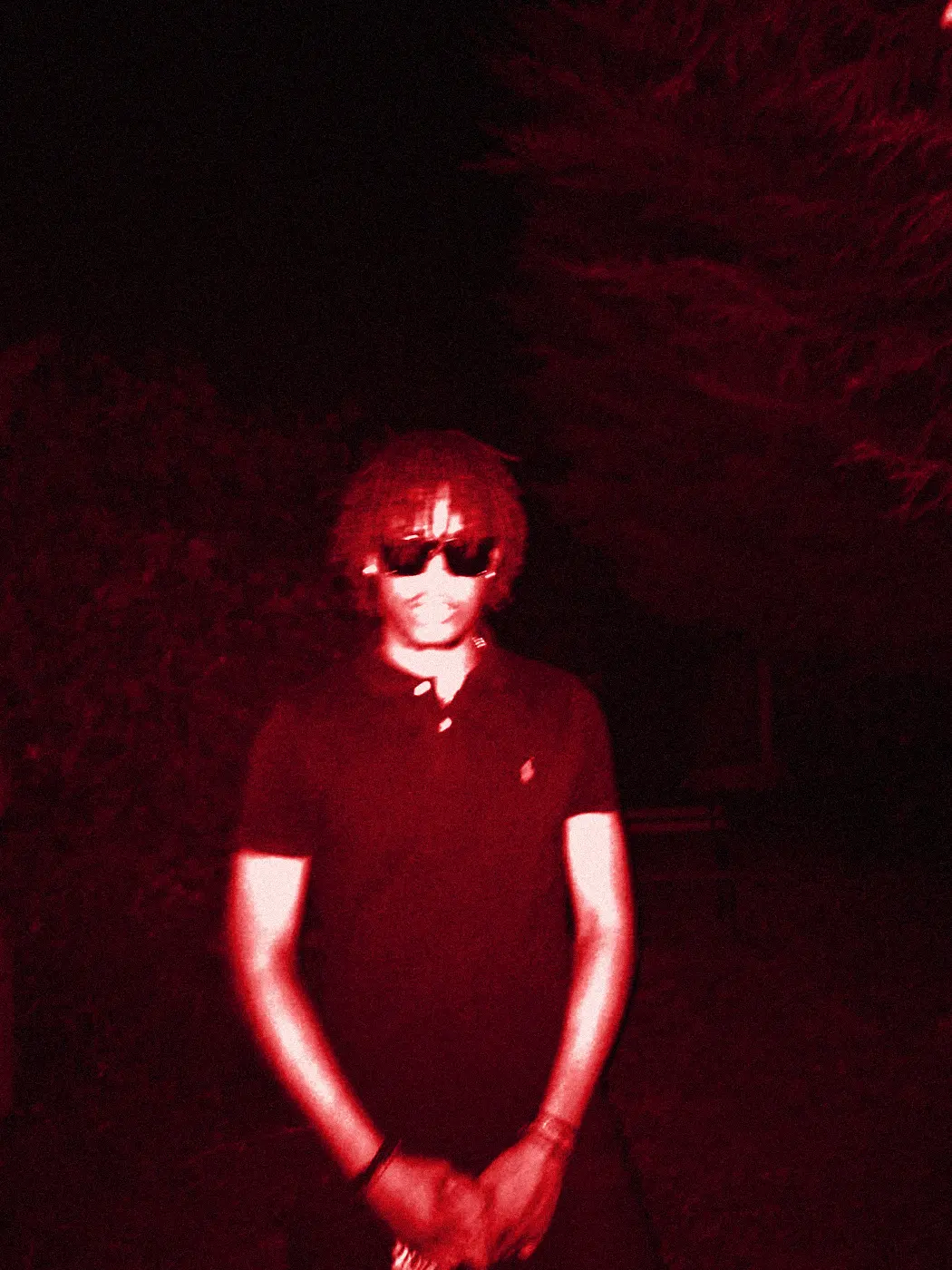 yukan in sunglasses, hands clasped, against dark hedges.